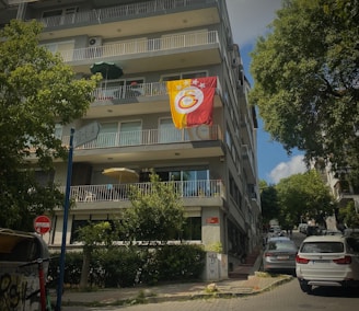 A multi-story residential building with a large flag displaying a sports emblem hanging from one of the balconies. The street is lined with trees, and several cars are parked alongside the building. A street sign and urban elements like a trash bin can be seen, creating a typical city neighborhood feel.