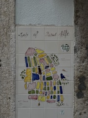 A decorative tiled map, titled 'Map of Bairro Alto', mounted on a textured wall. The map features various colored blocks and labels depicting different streets and areas. It includes artistic elements like a small drawing of a boat and some trees. The overall style is artistic and illustrative.
