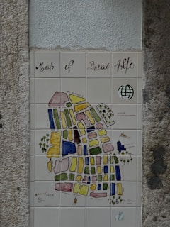 A decorative tiled map, titled 'Map of Bairro Alto', mounted on a textured wall. The map features various colored blocks and labels depicting different streets and areas. It includes artistic elements like a small drawing of a boat and some trees. The overall style is artistic and illustrative.