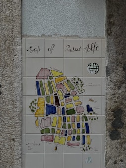 A decorative tiled map, titled 'Map of Bairro Alto', mounted on a textured wall. The map features various colored blocks and labels depicting different streets and areas. It includes artistic elements like a small drawing of a boat and some trees. The overall style is artistic and illustrative.