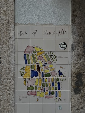 A decorative tiled map, titled 'Map of Bairro Alto', mounted on a textured wall. The map features various colored blocks and labels depicting different streets and areas. It includes artistic elements like a small drawing of a boat and some trees. The overall style is artistic and illustrative.