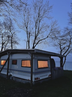 A caravan parked under a wooden shelter near a body of water during twilight. The windows are glowing with warm light, creating a cozy atmosphere. Bare trees with intricate branches surround the area, silhouetted against the fading light of the sky.