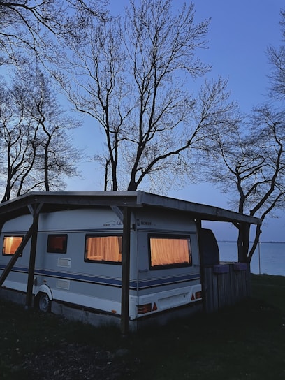 A caravan parked under a wooden shelter near a body of water during twilight. The windows are glowing with warm light, creating a cozy atmosphere. Bare trees with intricate branches surround the area, silhouetted against the fading light of the sky.