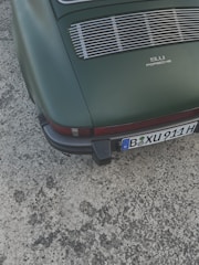 A vintage car with a rear view, featuring a matte green paint job and a classic 911 Porsche logo. The vehicle has a distinctive grille and a visible license plate at the bottom.