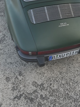 A vintage car with a rear view, featuring a matte green paint job and a classic 911 Porsche logo. The vehicle has a distinctive grille and a visible license plate at the bottom.