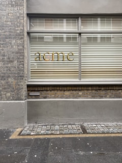 A brick wall with a window that has horizontal blinds partially closed. The word 'acme' is displayed in gold lettering on the glass. The facade includes a lower brick section and a pavement with a textured grid pattern adjacent to it.