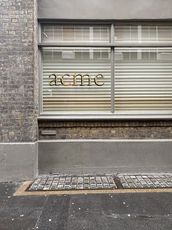 A brick wall with a window that has horizontal blinds partially closed. The word 'acme' is displayed in gold lettering on the glass. The facade includes a lower brick section and a pavement with a textured grid pattern adjacent to it.