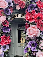 A doorway beautifully decorated with large, colorful artificial flowers. The flowers feature vibrant pinks, purples, and reds, arranged in a lush, arching display around the door, creating a whimsical and inviting entrance.