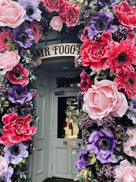 A doorway beautifully decorated with large, colorful artificial flowers. The flowers feature vibrant pinks, purples, and reds, arranged in a lush, arching display around the door, creating a whimsical and inviting entrance.