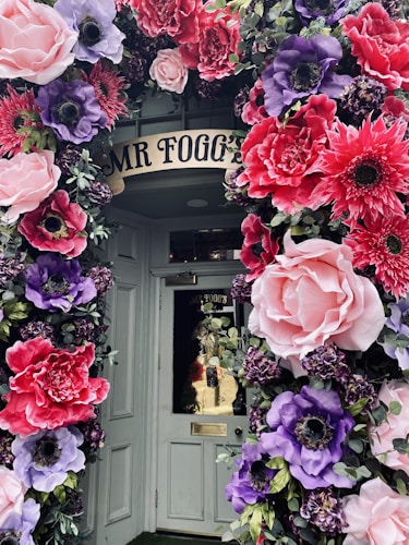 A doorway beautifully decorated with large, colorful artificial flowers. The flowers feature vibrant pinks, purples, and reds, arranged in a lush, arching display around the door, creating a whimsical and inviting entrance.