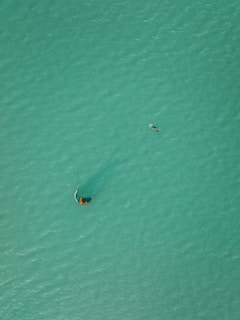 Aerial view of a person kite surfing on a vast expanse of turquoise water. The kite is visible above, creating a subtle ripple pattern around the surfer, who is gliding across the ocean surface. The image captures a sense of scale with the small figure against the large, uninterrupted area of water.