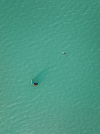 Aerial view of a person kite surfing on a vast expanse of turquoise water. The kite is visible above, creating a subtle ripple pattern around the surfer, who is gliding across the ocean surface. The image captures a sense of scale with the small figure against the large, uninterrupted area of water.