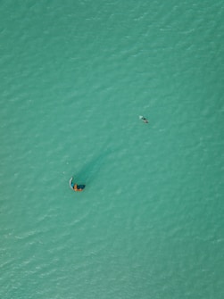 Aerial view of a person kite surfing on a vast expanse of turquoise water. The kite is visible above, creating a subtle ripple pattern around the surfer, who is gliding across the ocean surface. The image captures a sense of scale with the small figure against the large, uninterrupted area of water.