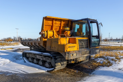 A large, yellow tracked vehicle is parked on snowy ground. The vehicle appears to be a construction or industrial machine with a spacious cabin. Surrounding the vehicle, the landscape is open with some snow cover on the ground, sparse vegetation, and there are a few utility poles in the distance.