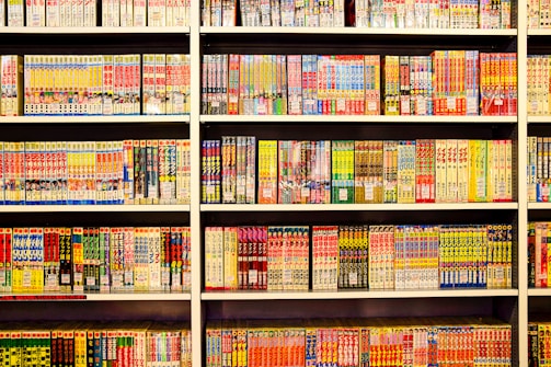 A bookshelf filled with numerous manga volumes organized neatly in rows. Each book is vividly colored, featuring different styles of artwork typical of Japanese manga. The spines display a variety of titles, characters, and symbols, creating a lively and engaging visual experience.