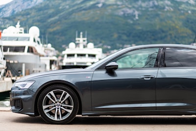 A sleek, modern car is parked by a marina, with several large yachts visible in the background. The scene is set against a backdrop of green hills and a cloudy sky, creating a luxurious and serene atmosphere.