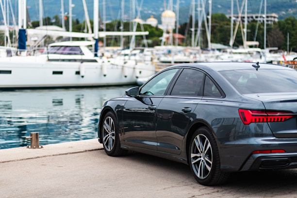 A sleek dark gray sedan is parked by a marina, where several white yachts are moored. The setting is picturesque, with the calm water reflecting the boats and the background of lush greenery and distant mountains.