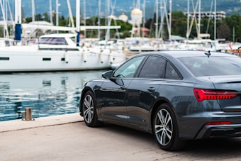 A sleek dark gray sedan is parked by a marina, where several white yachts are moored. The setting is picturesque, with the calm water reflecting the boats and the background of lush greenery and distant mountains.