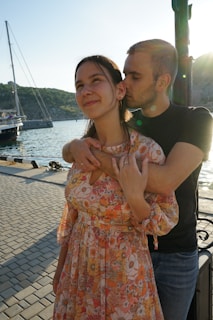 A couple stands on a waterfront; the woman in a floral dress is smiling slightly while the man, in a black shirt, hugs her from behind and kisses her cheek. A sailboat and the sea are visible in the background under a bright sky.