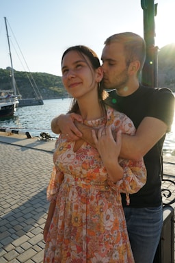 A couple stands on a waterfront; the woman in a floral dress is smiling slightly while the man, in a black shirt, hugs her from behind and kisses her cheek. A sailboat and the sea are visible in the background under a bright sky.