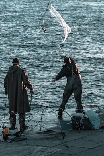 Two people dressed in fishing gear are standing by a body of water, one of them in the process of casting a large fishing net into the air. They are surrounded by various fishing equipment and bags on the ground.