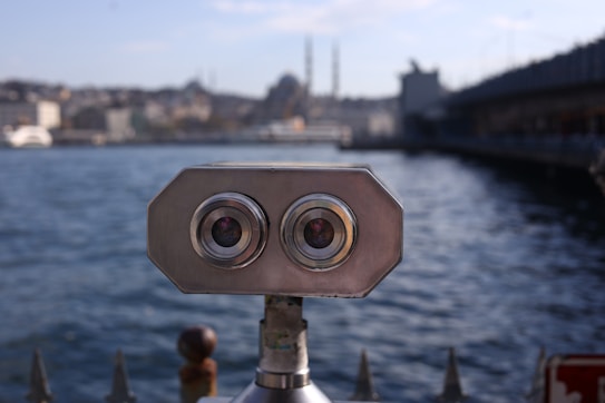 A public binocular device is positioned on a waterfront, with a body of water and an urban skyline in the background. The binoculars have a metallic finish and twin lenses. The scene includes buildings and a hazy sky.