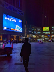 A nighttime urban scene featuring a large, brightly illuminated billboard displaying an advertisement with blue and white accents. A silhouette of a person stands in the foreground, holding a bag and looking toward the billboard. The area is a plaza with a few people and brightly colored illuminated vehicles. Surrounding buildings have a modern architecture style, with various lights and smaller advertisements visible.