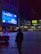 A nighttime urban scene featuring a large, brightly illuminated billboard displaying an advertisement with blue and white accents. A silhouette of a person stands in the foreground, holding a bag and looking toward the billboard. The area is a plaza with a few people and brightly colored illuminated vehicles. Surrounding buildings have a modern architecture style, with various lights and smaller advertisements visible.