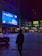 A nighttime urban scene featuring a large, brightly illuminated billboard displaying an advertisement with blue and white accents. A silhouette of a person stands in the foreground, holding a bag and looking toward the billboard. The area is a plaza with a few people and brightly colored illuminated vehicles. Surrounding buildings have a modern architecture style, with various lights and smaller advertisements visible.