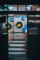 A laundromat interior featuring three large industrial washing machines, each with a digital interface and Japanese signage. The setting is illuminated with fluorescent lighting, and a blue bag is hanging on the side of one machine. The overall ambiance is a bit dim but functional, with visible steps leading up to the machines. The area appears clean and orderly, suggesting efficient use.