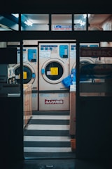 A laundromat interior featuring three large industrial washing machines, each with a digital interface and Japanese signage. The setting is illuminated with fluorescent lighting, and a blue bag is hanging on the side of one machine. The overall ambiance is a bit dim but functional, with visible steps leading up to the machines. The area appears clean and orderly, suggesting efficient use.