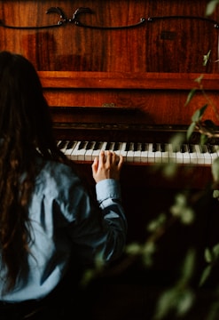 A person with long hair, dressed in a blue shirt, is playing a wooden piano. The setting is dimly lit, with the focus on a single hand touching the piano keys. Green foliage partially obscures the view, suggesting a natural, serene environment.