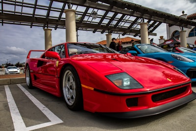 A bright red sports car is prominently parked, with sleek, aerodynamic design and distinctive branding details visible on the side. It is accompanied by a blue car to its right. The setting appears to be an outdoor car show or exhibition, with a modern structure and other cars in the background adding to the ambiance.