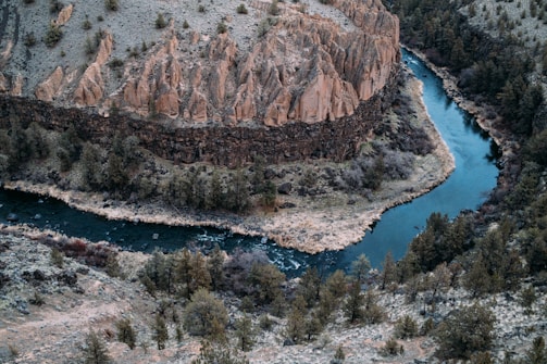 A winding river cuts through a rugged landscape featuring steep, rocky cliffs with jagged formations. The river is bordered by a variety of dense, green vegetation and trees, creating a vivid natural contrast. The terrain shows signs of erosion and weathering, hinting at the passage of time.