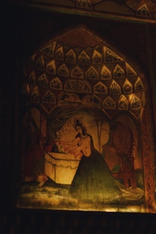 A detailed mural depicts a scene with two figures in traditional attire, surrounded by ornate patterns and decorative elements. The setting is reminiscent of a historical or cultural narrative, possibly a royal or ceremonial event, with intricate designs adorning the archway.