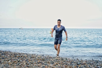 A man in a wetsuit is running out of the sea and onto a pebble beach. The ocean is calm, with a few small boats visible on the horizon under a cloudy sky.