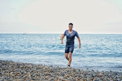 A man in a wetsuit is running out of the sea and onto a pebble beach. The ocean is calm, with a few small boats visible on the horizon under a cloudy sky.