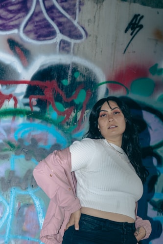 A person with long dark hair poses in front of a colorful graffiti-covered wall, wearing a white ribbed crop top with a pink jacket draped off the shoulders. The graffiti features abstract shapes and vibrant colors, including purple, green, red, and blue.