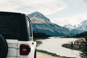 A rugged off-road vehicle is positioned in the foreground, overlooking a tranquil river meandering through a lush valley. Majestic snow-capped mountains loom under a partly cloudy sky, creating a breathtaking backdrop. Tall evergreen trees line the riverbanks, adding to the scenic beauty of the landscape.