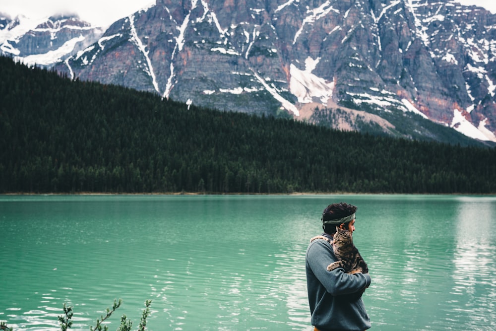 a man holding a cat standing next to a lake