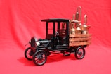 A vintage black model truck with intricate detailing and industrial equipment is positioned on a bright red background. The truck features a wooden flatbed holding several metallic distillation apparatuses, giving the scene a steampunk aesthetic.