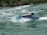 A blue jet boat labeled 'WHIRLPOOL JET' speeds through turbulent green waters, creating splashes and waves. The craft is partially covered with a glass section, housing seated passengers. Surrounding the river are lush green trees on rocky banks.