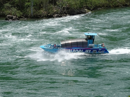 A blue jet boat labeled 'WHIRLPOOL JET' speeds through turbulent green waters, creating splashes and waves. The craft is partially covered with a glass section, housing seated passengers. Surrounding the river are lush green trees on rocky banks.
