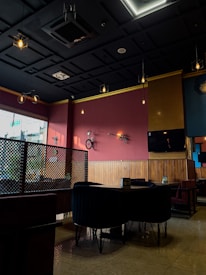 A modern, dimly lit cafe interior with maroon and wooden paneled walls. The ceiling features decorative hanging light bulbs. A bicycle and an electric guitar are mounted on the wall as decor. There are dark, plush seating arrangements and a lattice partition. A flat screen TV is mounted on the wall.