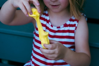 A child wearing a red and white striped sleeveless shirt is stretching a piece of bright yellow putty or slime between their hands. The background features a teal surface.