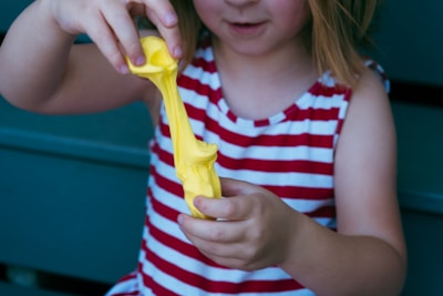 A child wearing a red and white striped sleeveless shirt is stretching a piece of bright yellow putty or slime between their hands. The background features a teal surface.