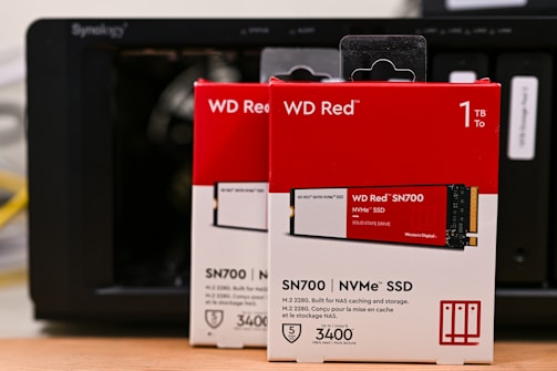 Boxes of WD Red SN700 NVMe SSDs, each with a storage capacity of 1TB, are positioned in front of a Synology NAS device. The boxes are predominantly red and white, featuring images of the SSDs and various specifications including read speed and warranty information.