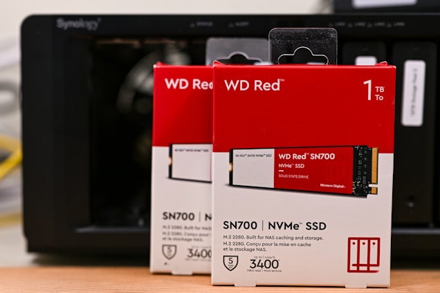 Boxes of WD Red SN700 NVMe SSDs, each with a storage capacity of 1TB, are positioned in front of a Synology NAS device. The boxes are predominantly red and white, featuring images of the SSDs and various specifications including read speed and warranty information.