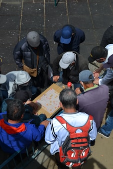 A group of people wearing caps and jackets are gathered around a table game, intently focused on playing. The scene is outdoors, and the players are crowded closely together, indicating a shared interest or enjoyment in the activity.