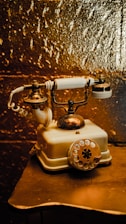 An antique rotary dial telephone with brass and ivory elements rests on a small table. The telephone is designed with intricate metalwork on the cradle and a coiled cord extending from the base. The background features a textured golden surface that adds a rich, warm hue to the scene.